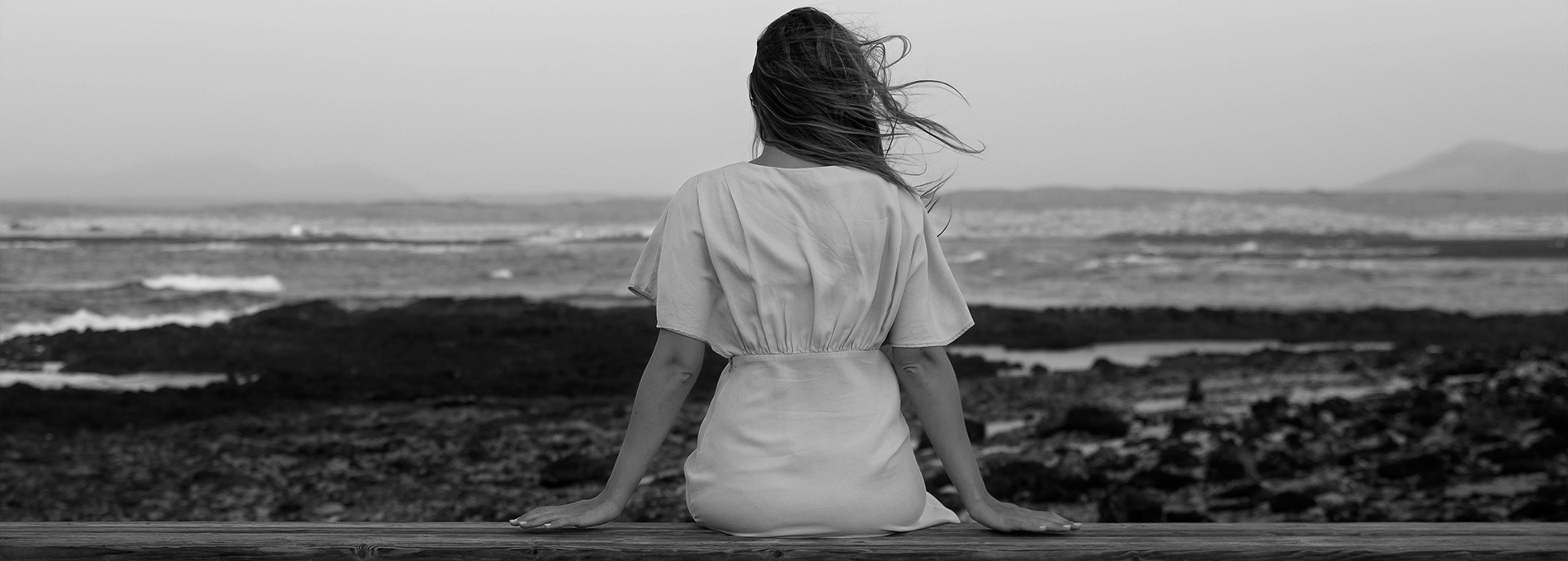 Black and white women sitting alone on bench looking out to sea with back to camera